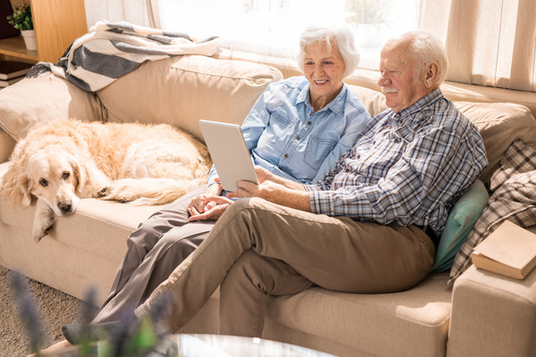 elderly m/f couple with dog on white couch looking at a tabl